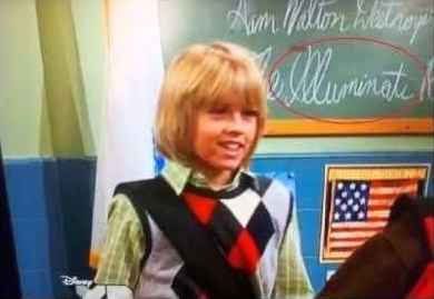 A boy stands in front of a chalkboard on which the word Illuminati is written